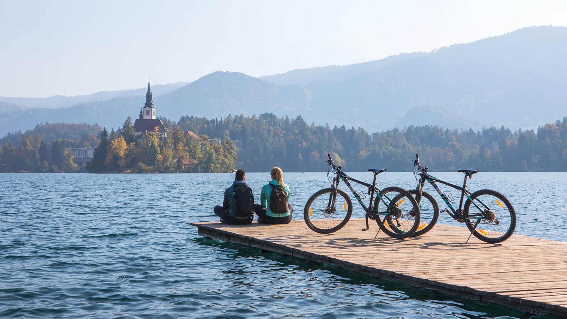 Zwei Personen sitzen auf einem Steg mit Fahrrädern und Blick auf See und Kirche (© Jost Gantar)