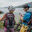 Zwei Radfahrer mit Helmen und Rucksäcken am See, Mann schaut auf sein Smartphone