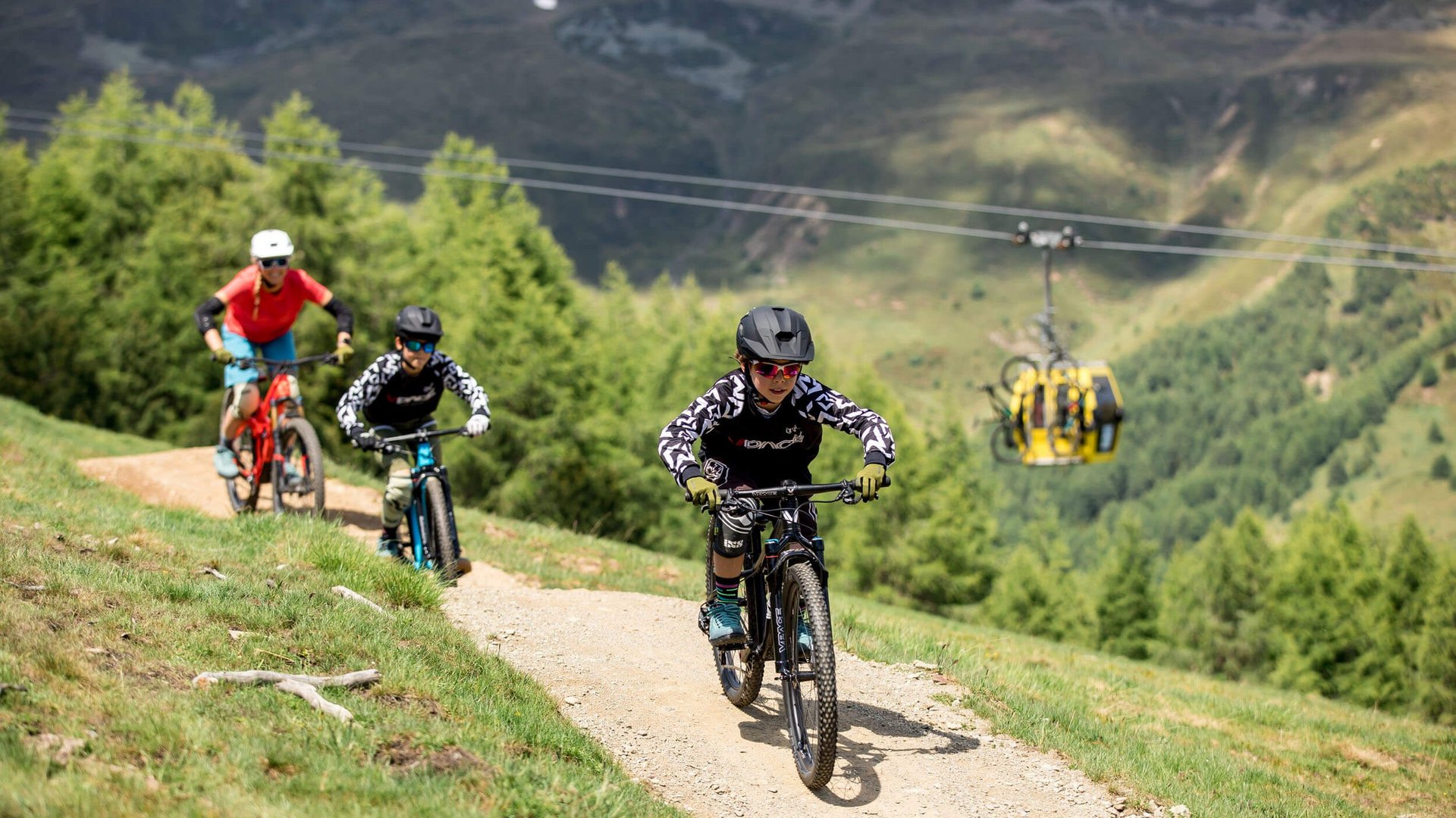 Drei Radfahrer fahren auf einem Bergweg mit Gondel im Hintergrund (© Anne Kaiser Photography)